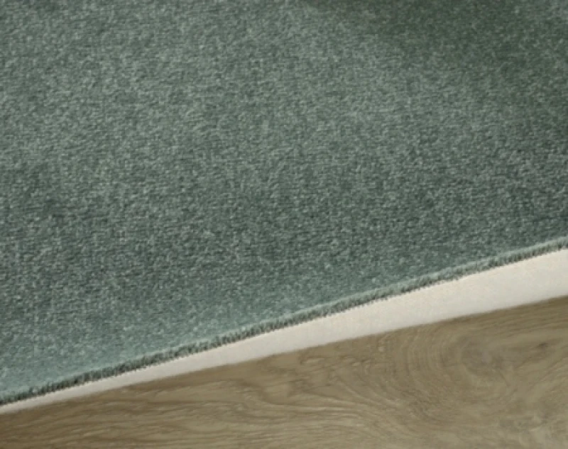 Close-up of a green textured carpet edge transitioning to a light wood floor, showing the beige underside and a diagonal seam.