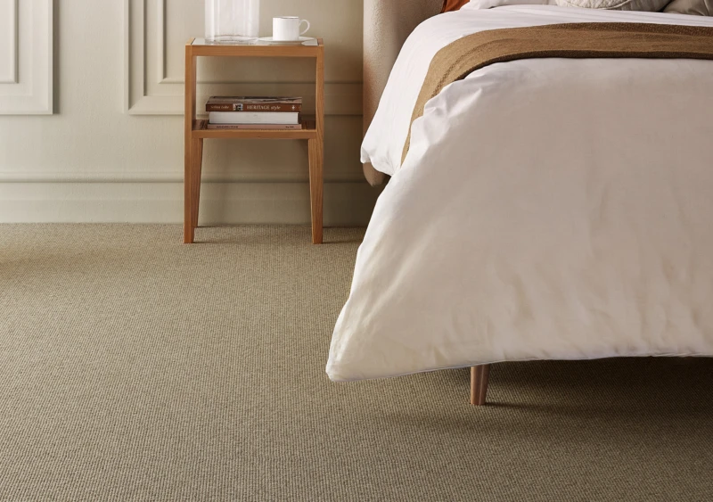 Wooden bedside table with a mug and books beside a bed with a white duvet on a beige carpet.