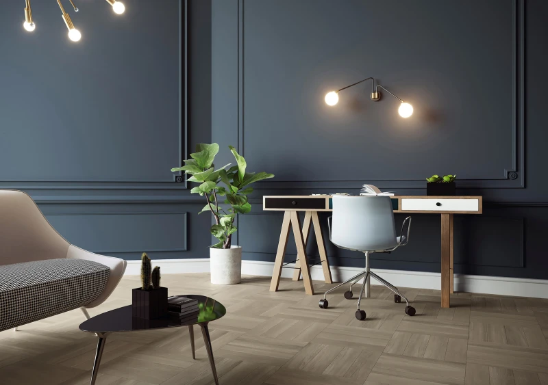 Modern home office with dark blue paneled walls, a wooden desk, light swivel chair, double-armed wall lamp, potted plant, and a curved beige sofa with a black glass coffee table.