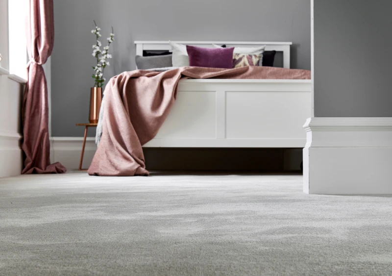 White platform bed with a pink throw and cushions against grey walls; pink curtain, and a copper vase with white flowers on a small side table.